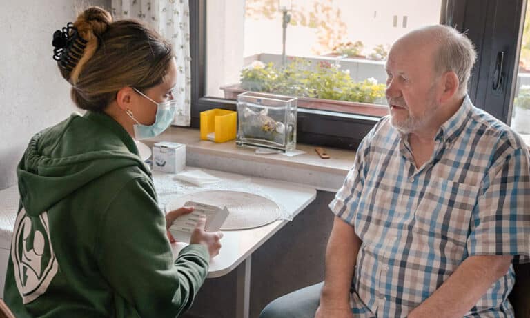 Eine Pflegekraft bespricht sich freundlich mit einem älteren Mann in kariertem Hemd.