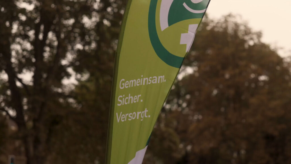 Grüne Flagge draußen, Text: „Gemeinsam. Sicher. Versorgt.“ Vor unscharfem Baumhintergrund.