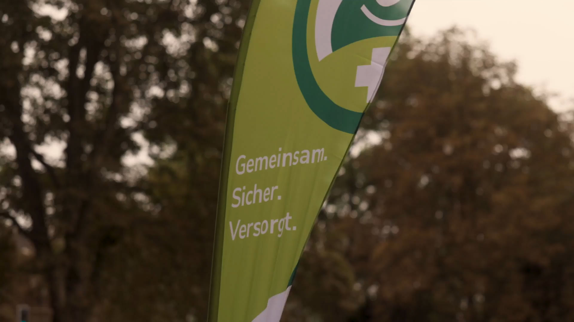 Grüne Flagge draußen, Text: „Gemeinsam. Sicher. Versorgt.“ Vor unscharfem Baumhintergrund.