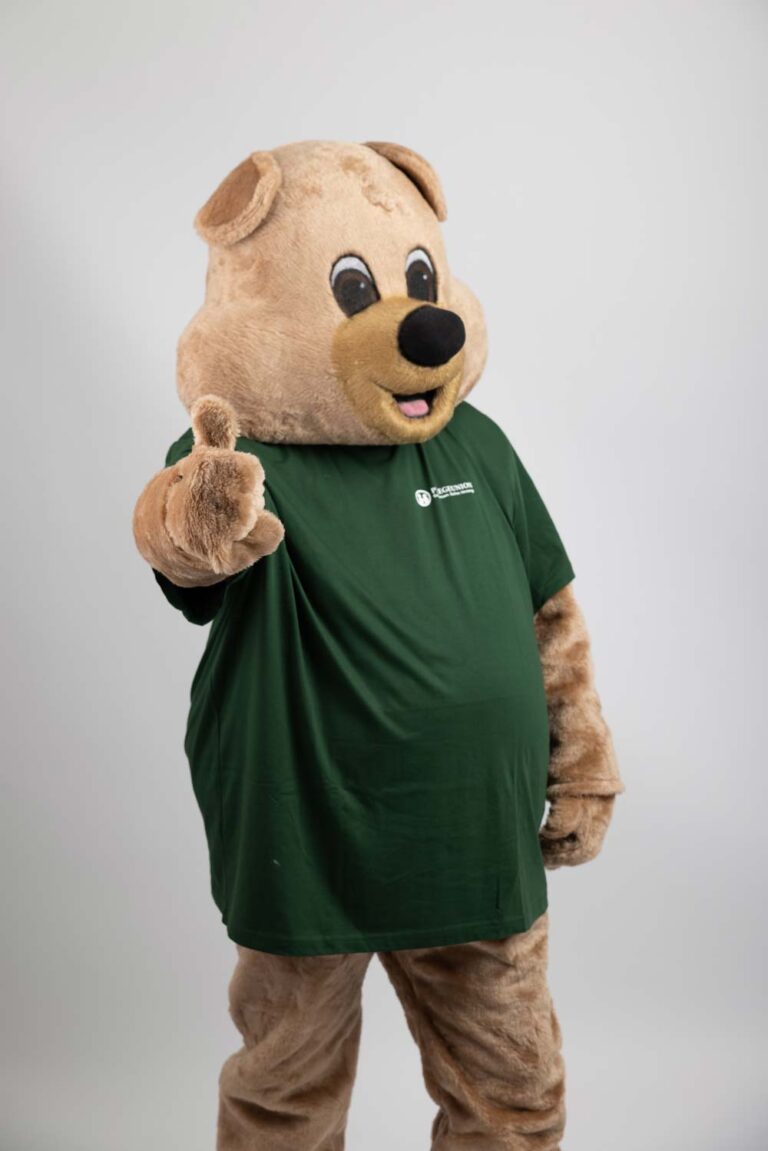 Ein lächelnder Mensch in einem beigen Bärenkostüm mit grünem T-Shirt, Daumen hoch.