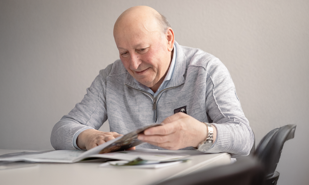 Ein älterer Mann, freundlich lächelnd, blättert in Zeitschriften, hellgrauer Pullover, helles Zimmer.
