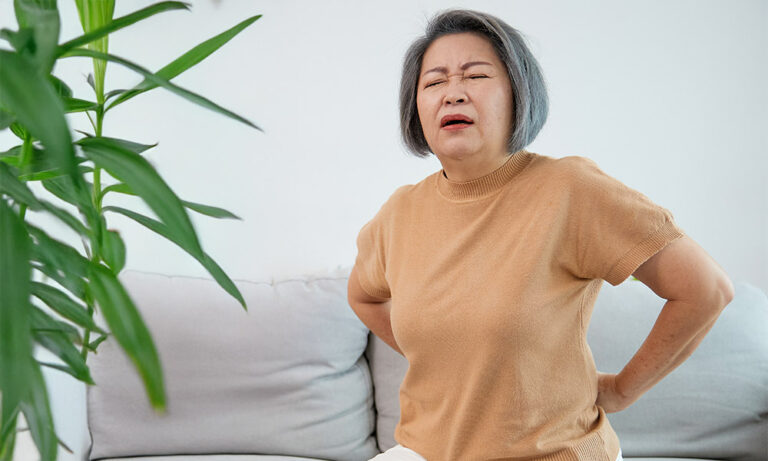 Eine ältere Dame mit Rückenschmerzen, beiger Pulli, sitzt auf einer Couch.