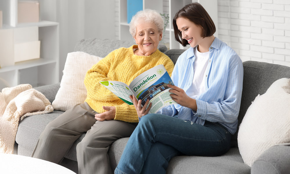 Ältere Dame und junge Frau, fröhlich auf grauem Sofa sitzend, lesen Magazin.