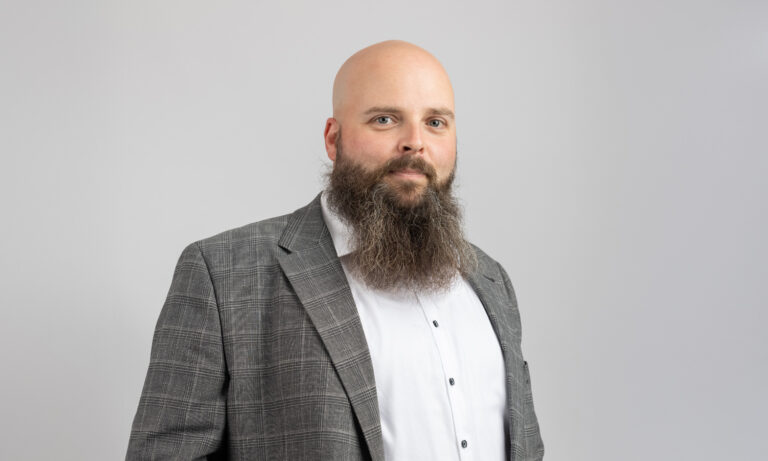 Ein glatzköpfiger Mann mit langem Bart trägt einen grauen Anzug und schaut ernst.