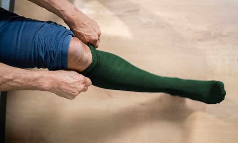 Eine Person zieht einen langen grünen Strumpf über das Knie hoch.