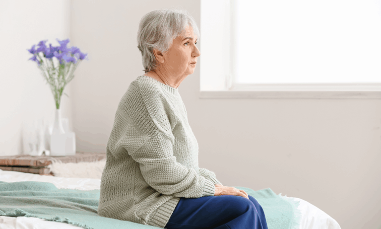 Ältere Frau im Profil, nachdenklich, grauer Pullover, blaue Hose, lila Blumen im Hintergrund.