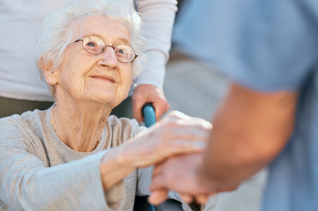 Händchenhaltende Pflegerin und ältere Frau im Rollstuhl zur Unterstützung im Freien im Altersheim. Liebe, Vertrauen und Gesundheitskrankenschwester oder medizinischer Wellness-Arzt für behinderte Patienten mit Freundlichkeit