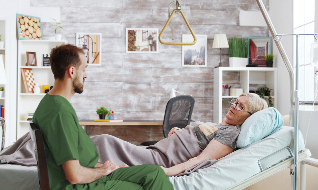 Ein Arzt in grüner Dienstkleidung sitzt neben einer älteren Frau im Krankenbett.