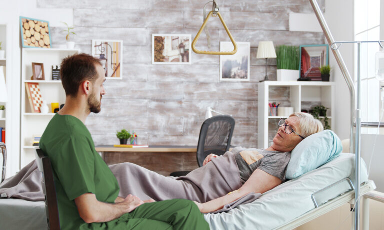 Ein Arzt in grüner Dienstkleidung sitzt neben einer älteren Frau im Krankenbett.