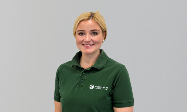 Eine lächelnde Person mit blonden Haaren in einem grünen Poloshirt vor grauem Hintergrund