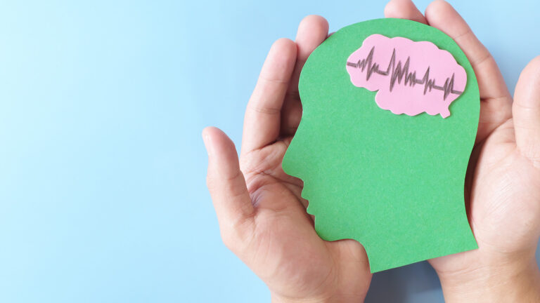 Zwei Hände halten eine grüne Kopfform mit rosa Gehirn und Herzlinien auf blauem Hintergrund