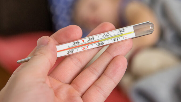 Eine Hand hält ein Fieberthermometer, im Hintergrund liegt unscharf ein Kind